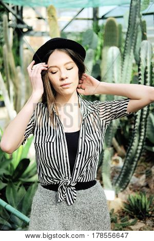 Young fashionable caucasian woman with long hair wearing hat and striped blouse basking in sunlight inside greenhouse near agave and cactus plants