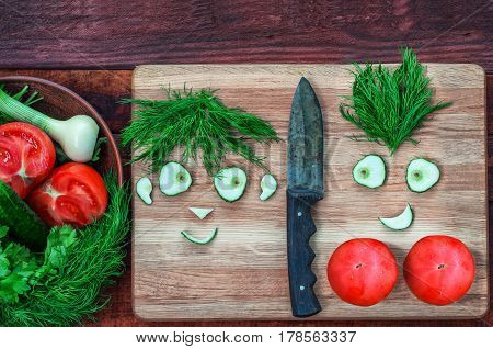 Fresh tomatoes and cucumber in a bowl and on a kitchen board funny faces males and women from pieces of cucumber and a bunch of dill top view