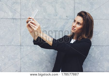 Portrait young businesswomen in black suit using smartphone isolated on background concrete gray wall mockup pretty hipster manager holding mobile gadget girl making selfie photo connect