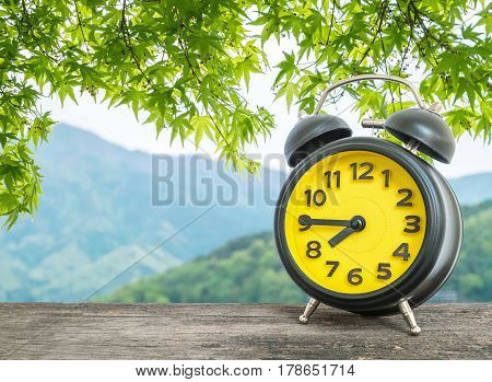 Closeup black and yellow alarm clock for decorate show a quarter to eight o c'clock or 7:45 a.m. on blurred leaves and mountain view background