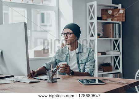 Concentraiting on project. Handsome young modern man holding a cup and looking at monitor while sitting at his working place in creative office