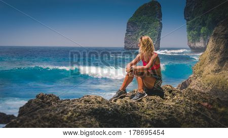 Girl sitting on the Rock and watching Huge Waves hitting Tembeling Coastline at Nusa Penida Island, Bali Indonesia.