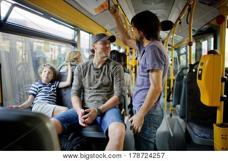 Passengers in the city bus. One man sits on a seat another stands nearby. They smile each other. At a window the boy sits. Modern public transport is convenient for children and adults.