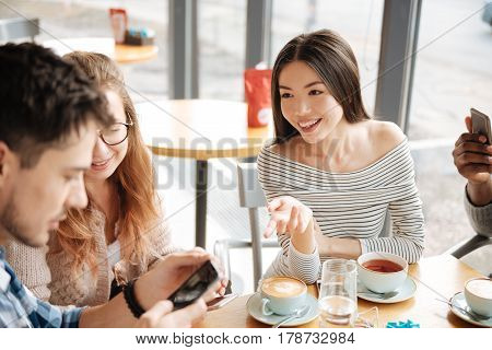 Discussing social media. Young smiling asian girl is gesting to her friends at cafe using smartphones.