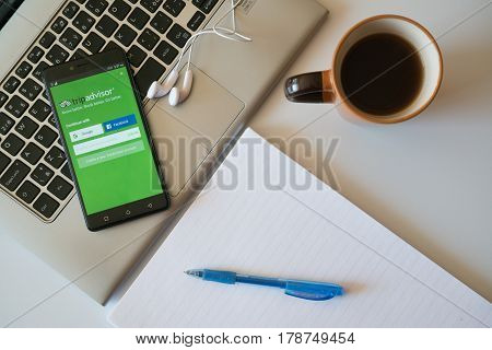 Nitra, Slovakia, march 28, 2017: Tripadvisor application in a mobile phone screen. Workplace with a laptop, an earphones, notepad and coffee.