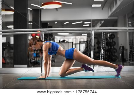 Attractive young woman is doing plank exercise while working out in gym. Beautiful slender brunette girl in blue sportswear.