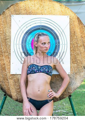 Attractive young smiling beautiful curly blonde hair slim girl fashion portrait in black swimming suit posing looking away calm look in target archery background.