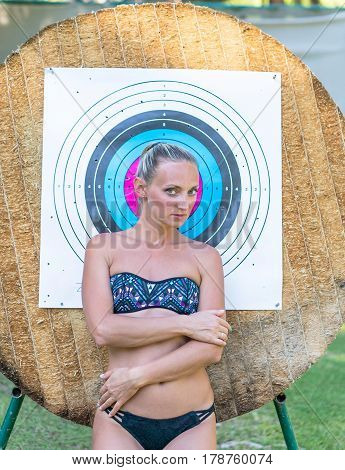 Attractive young smiling beautiful curly blonde hair slim girl fashion portrait in black swimming suit posing looking away calm look in target archery background.