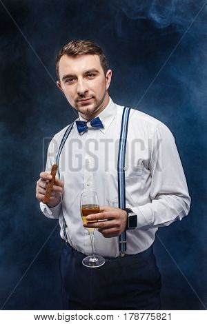 Portrait of a handsome man in bow-tie and suspenders holding glass of beverage and cigar while looking at camera. Smoke on background. Studio shot