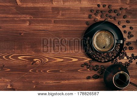 Hot coffee in black cup with crema beans and turkish pot cezve with copy space on brown old wooden board background top view. Rustic style.
