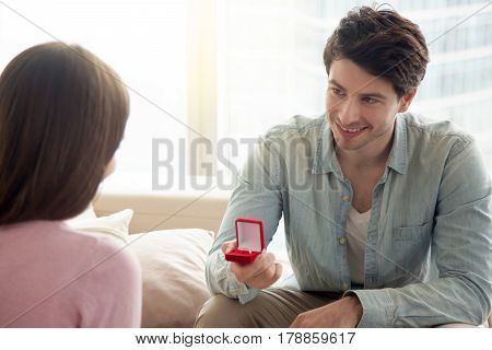 Young guy holding box with engagement ring, making marriage proposal to girlfriend, requesting a hand, indoors. Romantic concept, asking to marry, birthday gift, bride and groom wedding planning