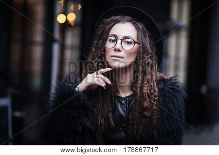 Portrait of a beautiful girl with brown eyes in glasses in a black hat and coat with fur in the cityscape looking in camera. The girl is like  Potter.
