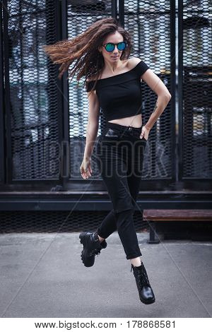 Full length portrait of young brunette girl in sunglasses jumping. Wearing checked shirt, blacg jeans and eyeglasses.