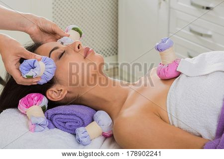 Herbal ball face massage in ayurveda spa. Female massagist with young woman in wellness center. Healthcare therapy to beautiful indian girl in beauty parlor, side view of face with eyes closed