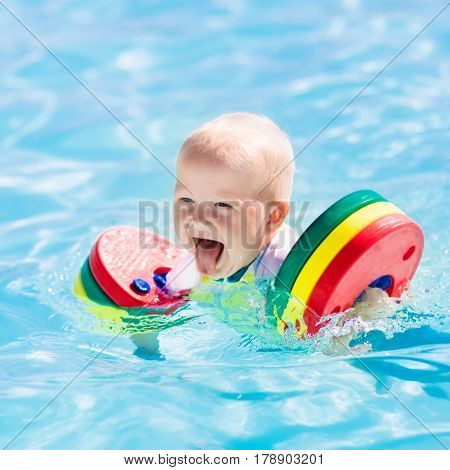Happy laughing little baby boy playing in outdoor swimming pool on a hot summer day. Kids learn to swim. Child with colorful floaties. Swimming aid for kid. Family vacation in tropical resort.