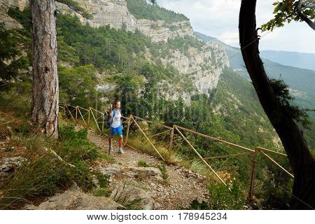 Young woman hiker with backpack walking a trail in rocky mountains with a stick in her hand. Magnificent landscape on background,