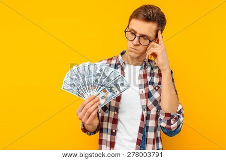 Pensive Man With Glasses Holding Dollar Bills In His Hands And Looking At Them On A Yellow Backgroun