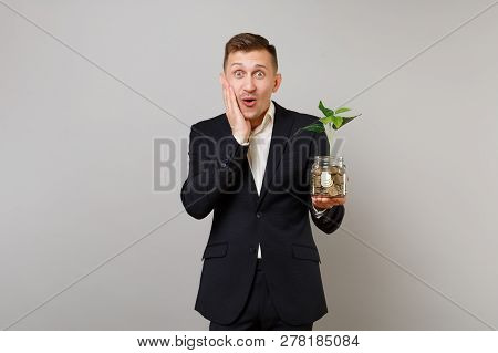 Surprised Young Business Man Put Hand On Cheek Holding Pile Gold Coins In Glass Jar With Green Plant