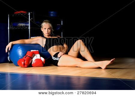 Bella combattente di MMA, seduta sul pavimento palestra con lei guanti e reclinata contro un pesante borsa afte