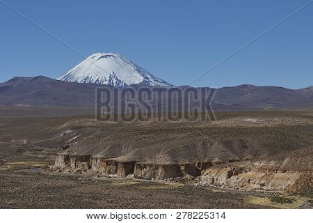 Snow Covered Parinacota Volcano (6342m) Towering Above The Altiplano And Cliffs Running Along The Va