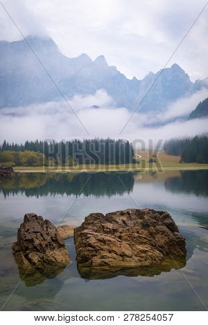 View Over Lake Laghi Di Fusine On A Foggy Morning With Some Rocks At The Beach And Mountain Range Ma