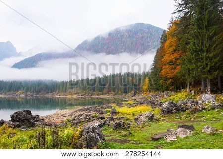 View Over Lake Laghi Di Fusine On A Foggy Morning With Some Rocks At The Beach And Mountain Range Ma