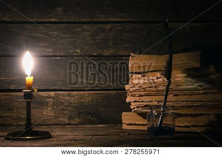 Quill Pen In A Inkpot And Stack Of Books In The Light Of Burning Candle On The Writer Desk Backgroun