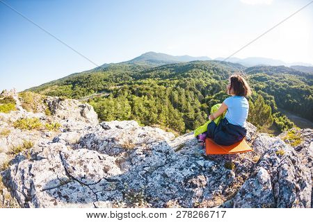 A Woman Is Sitting In A Sleeping Bag At Sunset. Camping On Top Of A Mountain. A Smiling Girl Rests A
