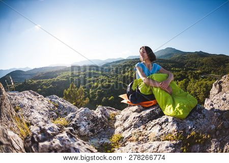 A Woman Is Sitting In A Sleeping Bag At Sunset. Camping On Top Of A Mountain. A Smiling Girl Rests A