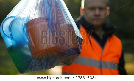 A Man In A Signal Orange Vest Holds A Package With Garbage On The Background Of Nature, Forest, Clos