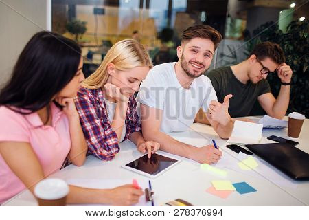 Handsome Young Bearded Man Sit Among His Coworkers And Hold Big Thumb Up. He Smiles On Camera. Other