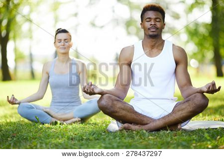 Calm concentrated young multi-ethnic yoga students sitting with crossed legs on grass and touching fingers in mudra while meditating in summer park