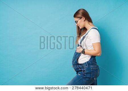 Pregnant Woman Caressing Her Belly Over Blue Background. A Cute Young Woman Wearing Glasses, And Den