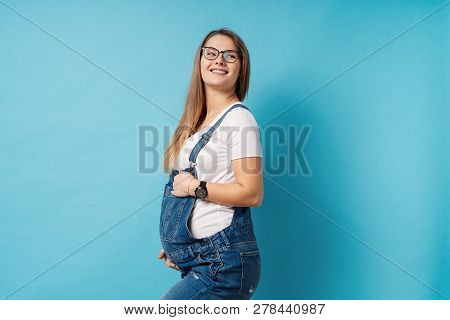 Pregnant Woman Caressing Her Belly Over Blue Background. A Cute Young Woman Wearing Glasses, And Den
