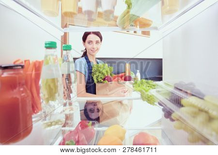 Portrait of female standing near open fridge full of healthy food, vegetables and fruits. Portrait of female
