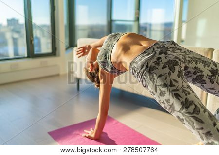 Yoga training at home - woman doing wild thing backbend pose in living room of apartment on exercise mat. View on sunset background with windows. Fitness active lifestyle.