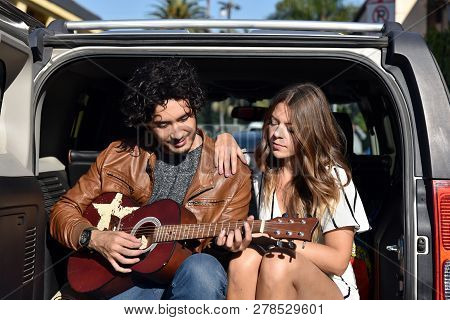Handsome Young Man Playing The Guitar For A Girl On The Tailgate Of His Car