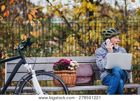 Senior Man With Electrobike Sitting On A Bench Outdoors In Town, Using Laptop And Smartphone.
