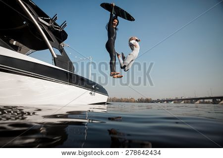 Two Blone And Brunette Girls Jumping To The River With Wakeboard On The Background Of Riverside