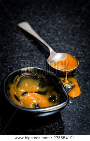 Caramel sauce in black dish and spilling from spoon over dark marble surface.