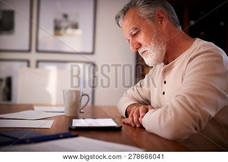 Senior Hispanic man sitting at a table using a tablet computer  in the evening, close up, side view