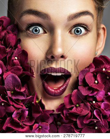 Closeup portrait of a surprised young woman. Female face with bright vivd makeup. Emotional girl with purple fowers.