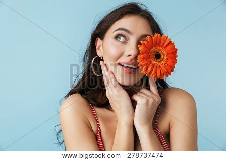 Lovely cheerful girl wearing summer clothes standing isolated over blue background, posing with a herbera flower