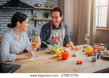 Cheerful Man Sit At Table With Wife In Kitchen. He Hold Piece Of Tomato On Fork And Show It To Woman
