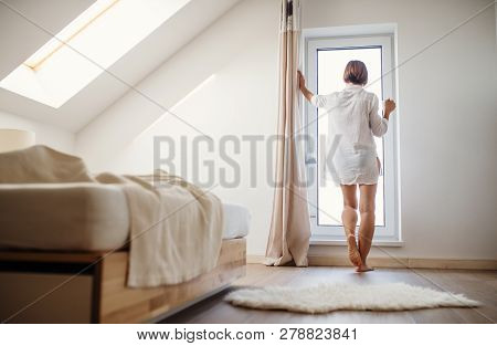 A Rear View Of Young Woman With Night Shirt Standing By The Window In The Morning.