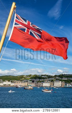 View of the waterfront with boats, the harbour and the historic buildings of Oban Scotland.  The Red Ensign flag is in the foreground, and McCaig's Tower can be seen in the distance.