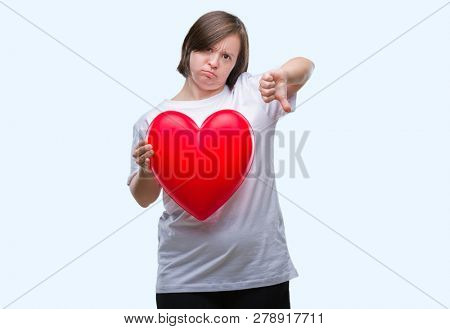 Young adult woman with down syndrome holding red heart over isolated background with angry face, negative sign showing dislike with thumbs down, rejection concept