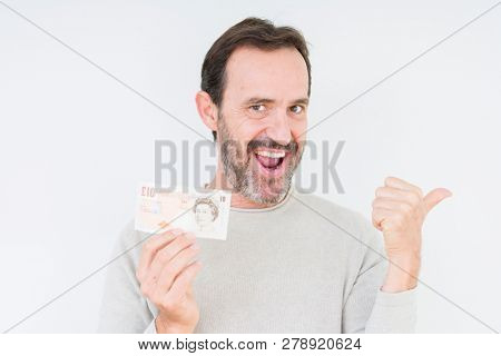 Senior man holding ten pounds bank note over isolated background pointing and showing with thumb up to the side with happy face smiling