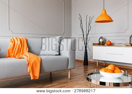 Orange Blanket On Grey Settee Next To Silver Table In Flat Interior With Lamp Above Cabinet. Real Ph