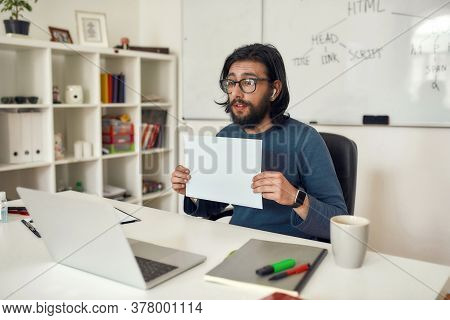 Young Male Computer Programming Tutor Wearing Eyeglasses Showing A Blank Sheet Of Paper At Web Camer